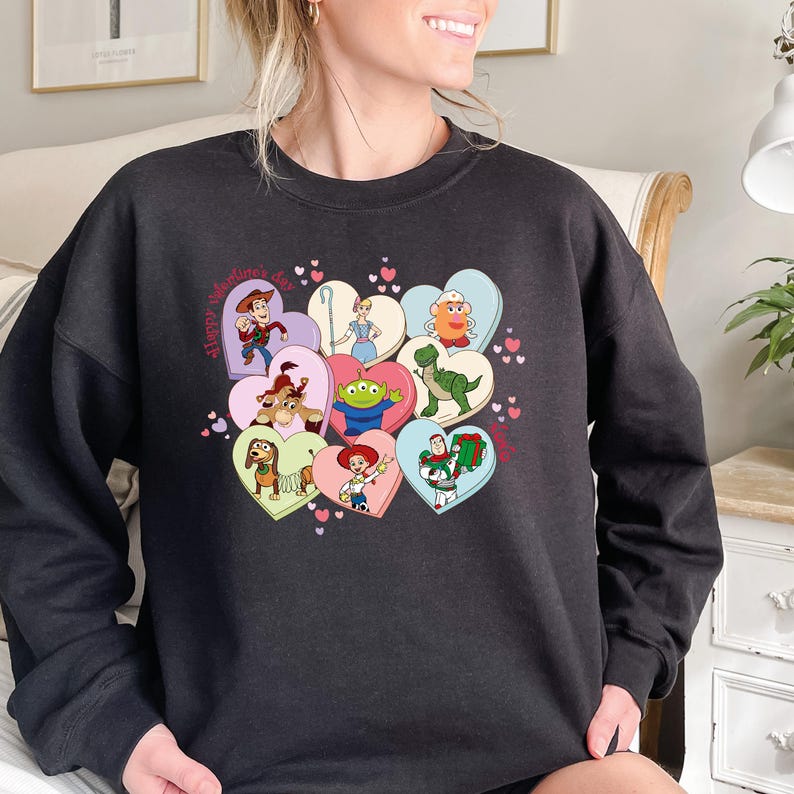 May include: Black sweatshirt featuring a colorful graphic of Toy Story characters inside heart-shaped designs. The phrase "Happy Valentine's Day" is printed on the left side. The design includes characters like Woody, Buzz Lightyear, and others.