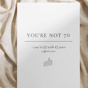 May include: White greeting card with the text "YOU'RE NOT 70" and the phrase "-you're 25 with 45 years experience". A small illustration of a cake with a lit candle is at the bottom. The card is on a beige fabric.