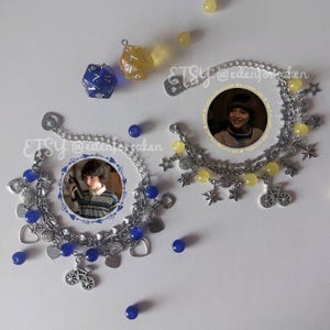 May include: Two silver charm bracelets with blue and yellow beads, heart and star charms, and bicycle pendants. Each bracelet features a circular image of a person. Scattered around are blue and yellow dice and beads.