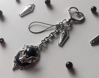 May include: A silver-toned keychain with a heart-shaped clasp, a chain, and a black bat pendant. The pendant has a black oval stone with a bat design. Additional coffin-shaped charms and black beads are scattered around.