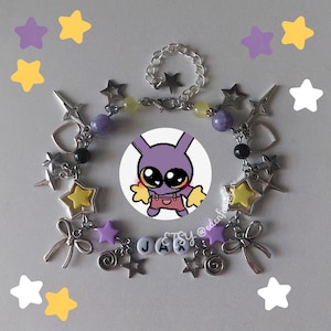 May include: A silver-toned charm bracelet featuring purple, yellow, and black beads, star-shaped charms, heart outlines, bows, and spiral charms. The bracelet includes white letter beads spelling JAX and a central circular graphic of a purple rabbit character.