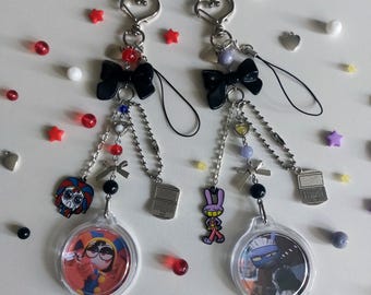 Op de afbeelding: Twee sleutelhangers met hartvormige sluitingen, zwarte strikken en bungelende kettingen. Elke sleutelhanger heeft een heldere, ronde bedel met een stripfiguur, een laptopbedel en diverse kralen in rood, wit en zwart. De achtergrond is wit met verspreide decoratieve elementen.