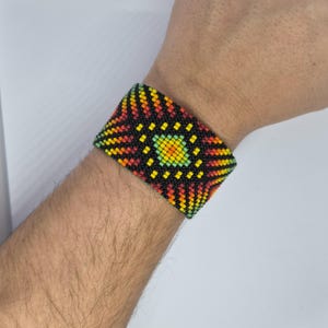 May include: A wide beaded bracelet with a geometric design. The bracelet has a central square pattern in yellow and green, with radiating lines in red, orange, and black. The bracelet is approximately 5 cm wide.