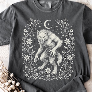 Snarling werewolf beneath a crescent moon framed by a floral wreath of wildflowers and trailing vines, in cream vintage folk-art linework on a pepper gray Comfort Colors tee.Snarling werewolf beneath a crescent moon framed by a floral wreath of wildflowers and trailing vines, in cream vintage folk-art linework on a pepper gray Comfort Colors tee.