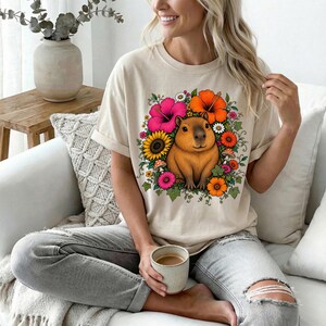 May include: A beige short-sleeve t-shirt featuring a central graphic of a brown capybara surrounded by a vibrant floral arrangement of pink, orange, and yellow flowers, green leaves, and small mushrooms, printed on the front of the garment.