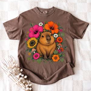 May include: A brown short-sleeve t-shirt featuring a central graphic of a capybara surrounded by colorful flowers including pink and orange hibiscus, a yellow sunflower, daisies, and small mushrooms with green leaves and vines.