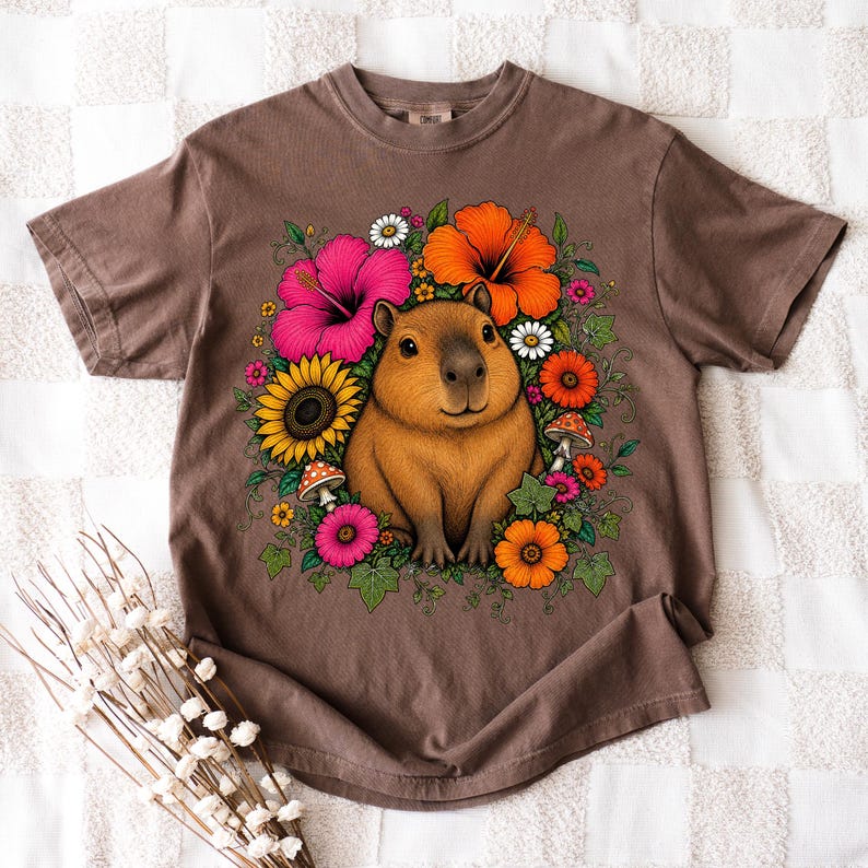 May include: A brown short-sleeve t-shirt featuring a central graphic of a capybara surrounded by colorful flowers including pink and orange hibiscus, a yellow sunflower, daisies, and small mushrooms with green leaves and vines.