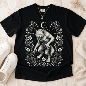 May include: A black short-sleeve t-shirt featuring a white graphic print of a snarling werewolf centered within an ornate floral frame. Above the creature is a crescent moon and small stars. The shirt has a crew neckline and a tag at the collar.