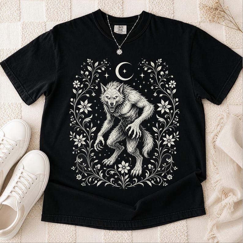 May include: A black short-sleeve t-shirt featuring a white graphic print of a snarling werewolf centered within an ornate floral frame. Above the creature is a crescent moon and small stars. The shirt has a crew neckline and a tag at the collar.