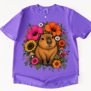 May include: A purple short-sleeve t-shirt featuring a central graphic of a brown capybara surrounded by a colorful arrangement of pink and orange hibiscus flowers, yellow sunflowers, small white daisies, green leaves, and red spotted mushrooms.