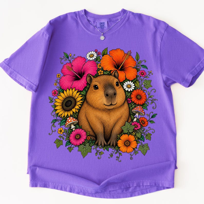May include: A purple short-sleeve t-shirt featuring a central graphic of a brown capybara surrounded by a colorful arrangement of pink and orange hibiscus flowers, yellow sunflowers, small white daisies, green leaves, and red spotted mushrooms.
