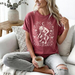 May include: A short-sleeved, crew-neck t-shirt in a muted red color. The front features a white graphic print of a werewolf standing on its hind legs, surrounded by a decorative frame of swirling vines, flowers, and a crescent moon at the top.