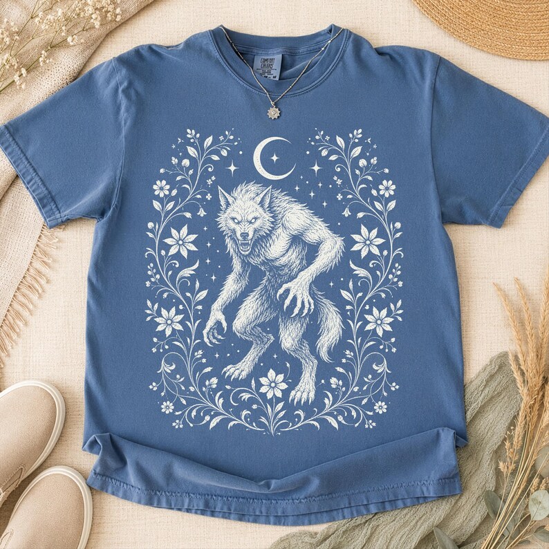 May include: A blue short-sleeve t-shirt featuring a white graphic print of a werewolf standing in the center, surrounded by a decorative floral frame and a crescent moon with stars above the creature. The shirt has a crew neckline and a relaxed fit.