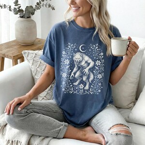 May include: A blue short-sleeve t-shirt featuring a white graphic print of a werewolf standing within a decorative frame of flowers, leaves, and a crescent moon. The shirt has a crew neckline and a relaxed fit, worn with light gray distressed jeans.