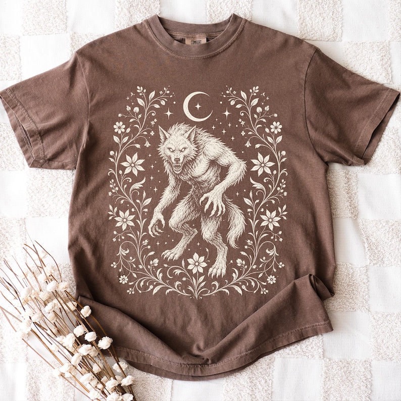 May include: A brown short-sleeve t-shirt featuring a cream-colored graphic print of a werewolf standing in the center. The design is framed by intricate floral vines and a crescent moon with small stars positioned above the creature's head.