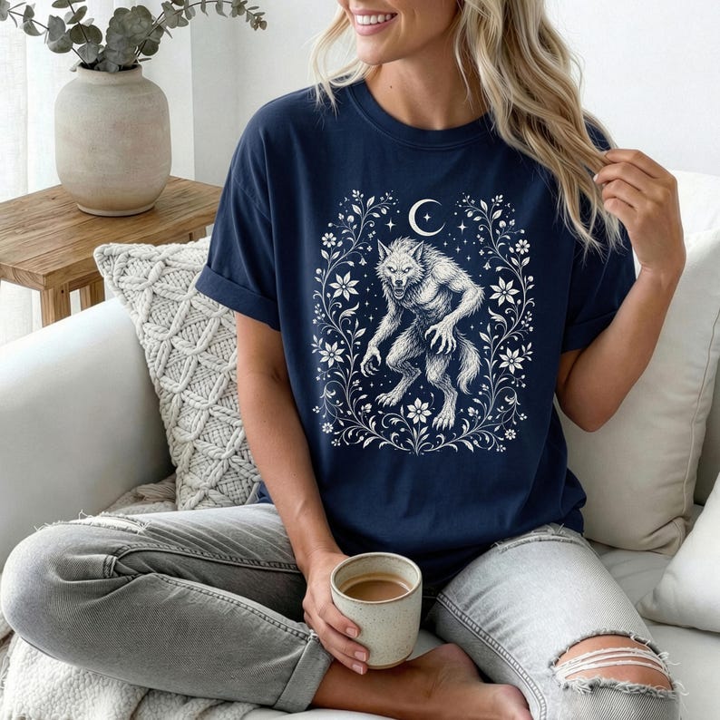 May include: A navy blue t-shirt featuring a white graphic print of a werewolf standing within a decorative frame of flowers, leaves, and a crescent moon. The shirt has short sleeves and a crew neckline, showcasing a detailed illustration.