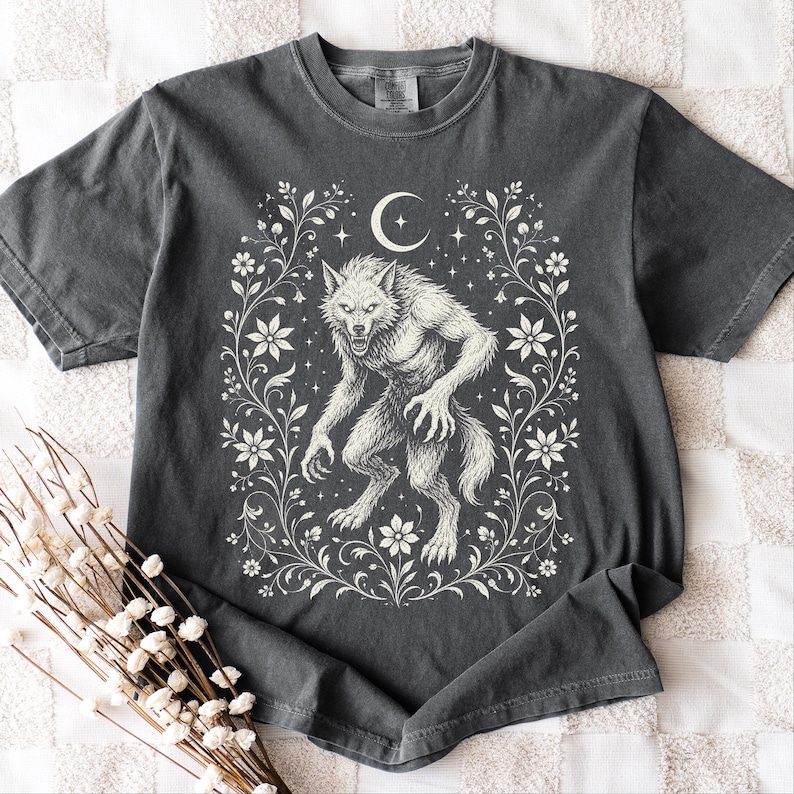 Snarling werewolf beneath a crescent moon framed by a floral wreath of wildflowers and trailing vines, in cream vintage folk-art linework on a pepper gray Comfort Colors tee.Snarling werewolf beneath a crescent moon framed by a floral wreath of wildflowers and trailing vines, in cream vintage folk-art linework on a pepper gray Comfort Colors tee.