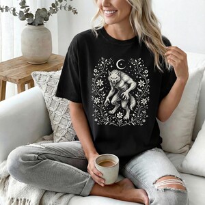 May include: A black short-sleeve t-shirt featuring a white graphic print of a werewolf standing in the center, surrounded by a detailed floral frame and a crescent moon at the top. The shirt is paired with light gray distressed denim jeans.