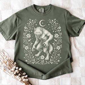May include: An olive green short-sleeve t-shirt featuring a cream-colored graphic print of a werewolf standing in the center. The design is framed by intricate floral vines, a crescent moon, and small stars, with a Comfort Colors brand label at the neck.