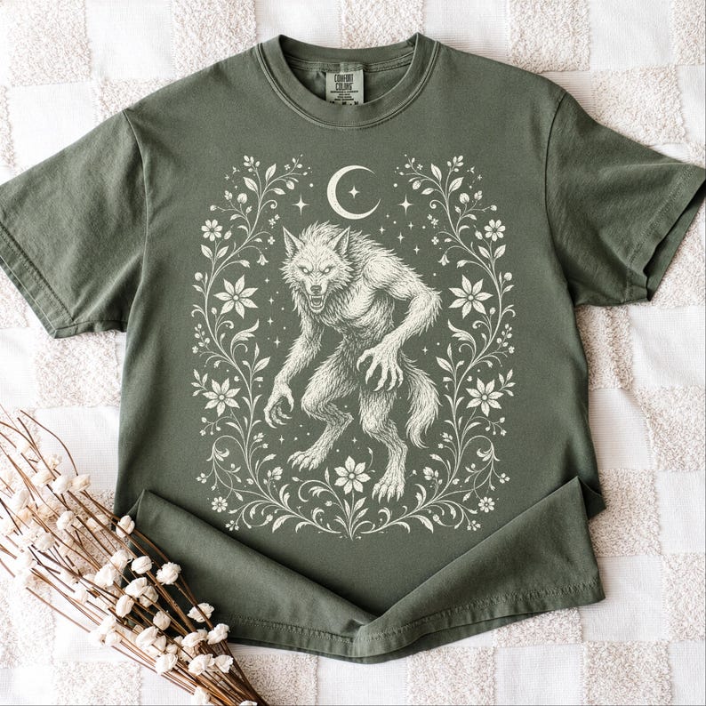 May include: An olive green short-sleeve t-shirt featuring a cream-colored graphic print of a werewolf standing in the center. The design is framed by intricate floral vines, a crescent moon, and small stars, with a Comfort Colors brand label at the neck.