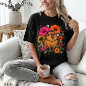 May include: A black short-sleeve t-shirt featuring a central graphic of a brown capybara surrounded by a vibrant arrangement of colorful flowers including pink hibiscus, orange blossoms, yellow sunflowers, and small red mushrooms with green leaves.