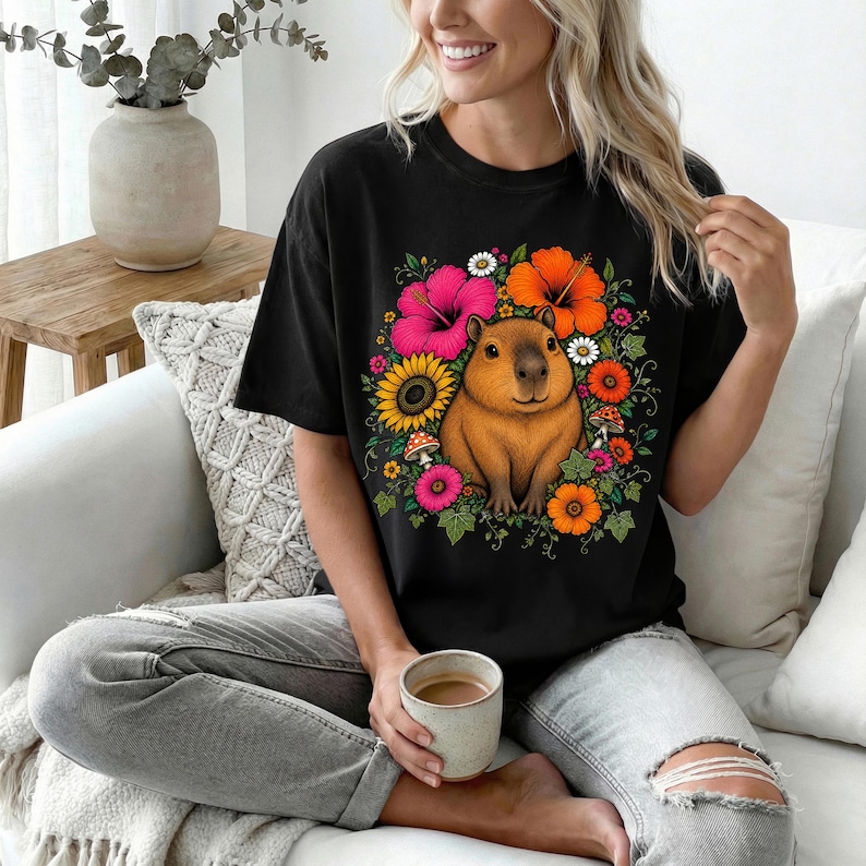 May include: A black short-sleeve t-shirt featuring a central graphic of a brown capybara surrounded by a vibrant arrangement of colorful flowers including pink hibiscus, orange blossoms, yellow sunflowers, and small red mushrooms with green leaves.