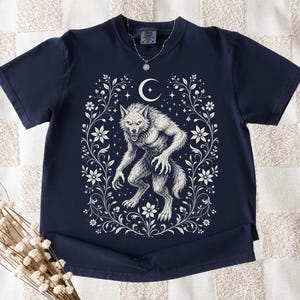 May include: A navy blue short-sleeve t-shirt featuring a white graphic print of a werewolf centered within an ornate floral frame. Above the werewolf is a crescent moon and small stars. A silver-toned necklace with a star pendant is visible.