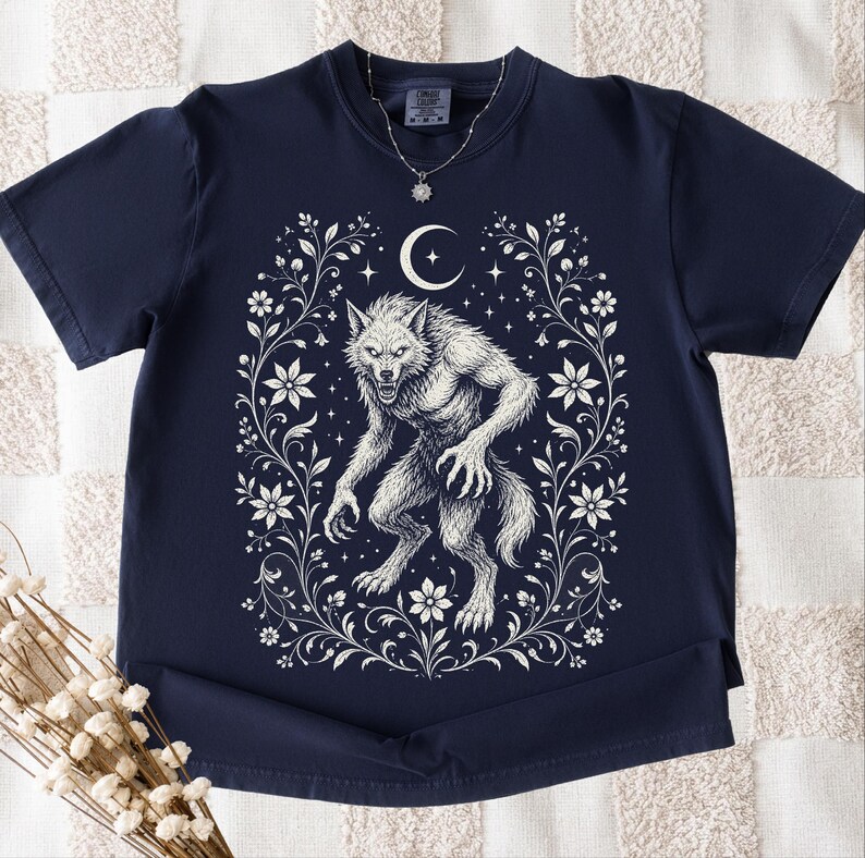 May include: A navy blue short-sleeve t-shirt featuring a white graphic print of a werewolf centered within an ornate floral frame. Above the werewolf is a crescent moon and small stars. A silver-toned necklace with a star pendant is visible.