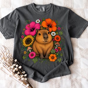 May include: A charcoal gray t-shirt featuring a central graphic of a brown capybara surrounded by a vibrant arrangement of colorful flowers including pink and orange hibiscus, yellow sunflowers, white daisies, and small red-spotted mushrooms.