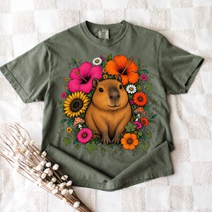 May include: An olive green short-sleeve t-shirt featuring a central graphic of a brown capybara surrounded by a vibrant arrangement of pink hibiscus, orange hibiscus, yellow sunflowers, small white daisies, and red mushrooms with green leaves.