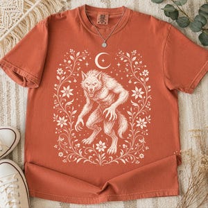 May include: A rust-colored short-sleeve t-shirt featuring a cream-colored graphic print of a werewolf centered within a decorative floral frame. Above the werewolf is a crescent moon and small stars. The shirt has a crew neckline and a tag.