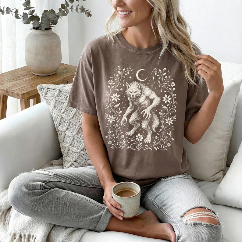 May include: A brown short-sleeve t-shirt featuring a cream-colored graphic print of a werewolf centered within an ornate floral frame topped with a crescent moon. The shirt has a crew neckline and a relaxed fit, paired with light blue distressed jeans.