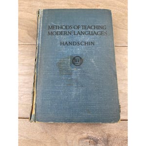 May include: A blue hardcover book titled Methods of Teaching Modern Languages by Handschin. The front cover features a small circular emblem with an open book icon in the center. The spine shows signs of wear with visible fraying and paper loss.