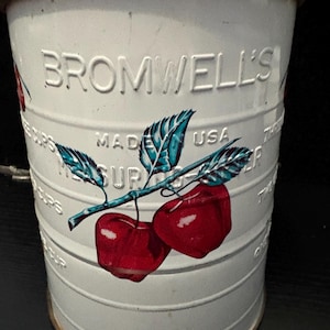 以下が含まれることがあります： 「BROMWELL'S」と「MADE IN USA」のエンボス加工が施された白い金属製の計量カップ。青と緑の葉が付いた2つの赤いサクランボの塗装デザインが特徴です。カップには測定マークが付いています。