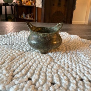 May include: A small, antique bronze-colored bowl with two handles. The bowl has a rounded body and a textured surface. It sits on a white, crocheted doily with a scalloped edge. The background includes a wooden table and antique furniture.