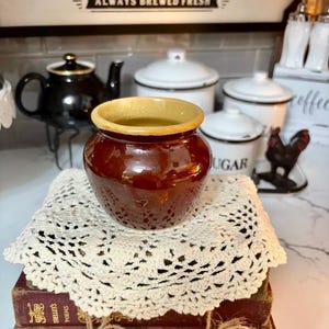 May include: A small, brown ceramic jar with a light tan rim sits atop a cream-colored, crocheted doily. The jar is placed on a stack of antique books. In the background are white canisters and a black teapot.