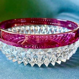 Puede incluir: Un cuenco redondo de cristal con un borde de color magenta intenso y un patrón prensado texturizado. La parte inferior del cuenco es de cristal transparente y muestra un patrón geométrico de corte de diamante pronunciado en el exterior.