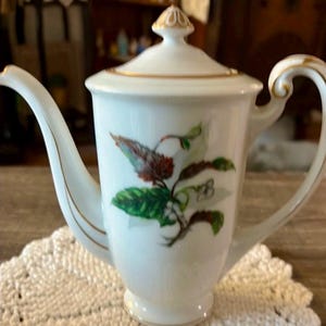 Pode incluir: Cafeteira de porcelana branca com tampa, com um desenho floral em verde, vermelho e castanho. A cafeteira tem um bico e uma pega curvos, com acabamento dourado. Está sobre uma toalha de crochet branca.