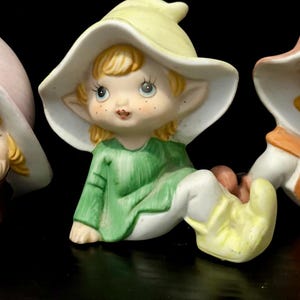 Könnte beinhalten: Keramikfigur eines sitzenden Elfen mit grünem Hemd, weißer Hose und gelben Schuhen. Der Elf hat einen gelben Hut mit weißer Krempe und blondem Haar. Die Figur hat ein bemaltes Gesicht mit blauen Augen und Sommersprossen.