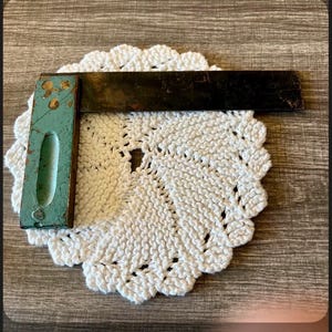 May include: An antique carpenter's square with a teal handle and a dark metal blade rests on a white, crocheted doily. The square shows signs of wear, adding to its vintage appeal. The doily has a scalloped edge.