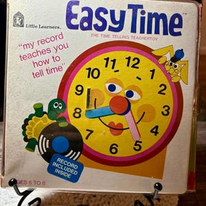 May include: Vintage "Easy Time" educational toy box. The box features a large, smiling clock face with a yellow background, pink and red border, and blue and pink clock hands. The box also includes a record and a cartoon rabbit.