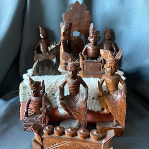 Puede incluir: Una colección de figuras de madera talladas a mano que representan músicos y bailarines con vestimenta tradicional, incluyendo un pequeño instrumento de madera con cinco componentes circulares, sobre un paño de ganchillo blanco y libros.