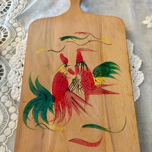 May include: Wooden cutting board with a handle, featuring a hand-painted design of two roosters. The roosters are primarily red with green and yellow accents. The board is rectangular with a rounded top and a hole for hanging.