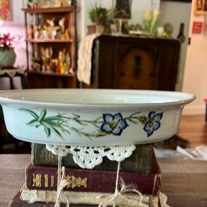 May include: White oval serving dish with a floral design featuring blue flowers and green leaves. The dish is resting on a stack of vintage books tied with twine and a white lace doily. A decorative piece for the home.