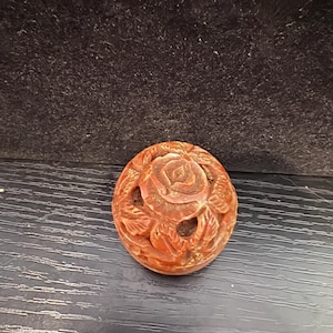 Peut inclure: Un objet rond sculpté, de couleur brun rougeâtre, représentant une rose. La rose est au centre, entourée de feuilles sculptées et d'une bordure circulaire. L'objet est probablement en pierre ou dans un matériau similaire.