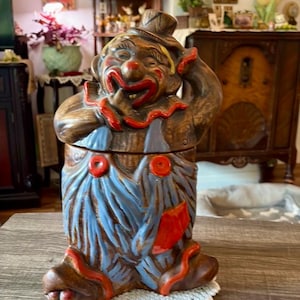 Könnte beinhalten: Ein Vintage-Clown-Keksdose aus Keramik. Die Dose ist braun mit blauen Latzhosen, roten Akzenten und einem lächelnden Gesicht. Der Clown trägt einen Hut und hat eine rote Nase und einen roten Mund. Die Dose steht auf einer weißen Spitze.