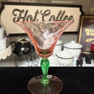 May include: A stemmed glass with a wide, pink, textured bowl featuring a rippled surface. The stem is bright green with a twisted, spiral pattern, and the circular base is clear pink glass. The glass stands upright on a flat surface.