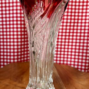 May include: A clear glass vase featuring a deep red rim and a scalloped, wavy top edge. The body of the vase is decorated with vertical, textured, leaf-like patterns that extend from the base to the top, creating a light-reflecting, faceted surface.