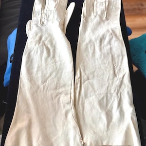 May include: A pair of long, off-white leather gloves featuring a smooth, matte finish and a classic five-finger design. The gloves extend past the wrist, showcasing a structured construction with visible stitching along the fingers and seams.
