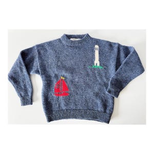 May include: A blue knit sweater with a crew neck and long sleeves. The sweater features a red sailboat and a white lighthouse with green grass. The sweater has ribbed cuffs and hem.